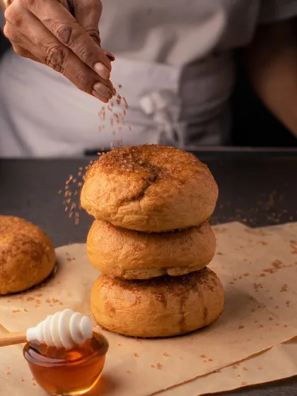 A baker sprinkling brown sugar over a stack of fresh bagels. This action shot captures the final touch that makes the food special.