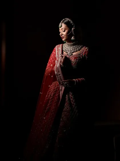 A full-length portrait in shadow, creating a dramatic and artistic feel. The focus remains on the bride's silhouette and the richness of her red lehenga.