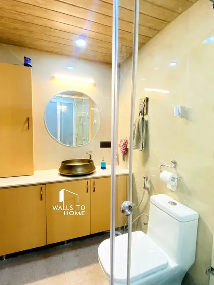 A bright and functional bathroom with light-colored tiles and wooden-finish cabinets. The design includes a round mirror, a stylish brass-colored vessel sink, and a glass partition for the shower area.