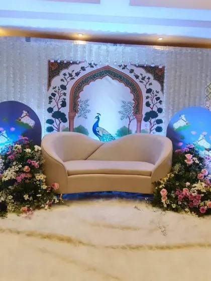 A beautiful stage decoration with a royal Indian theme. The backdrop features peacock and palace art, with a curved sofa and floral arrangements creating a regal seating area.
