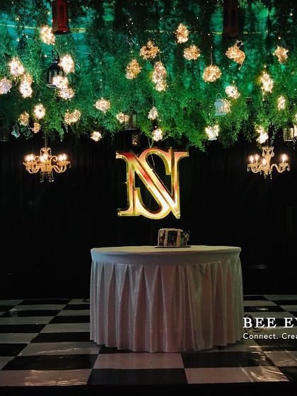 A chic cocktail event setup featuring a custom monogram and a ceiling adorned with hanging greenery and lights. The checkered dance floor adds a classic touch, making it the perfect space for a night of celebration and dancing.