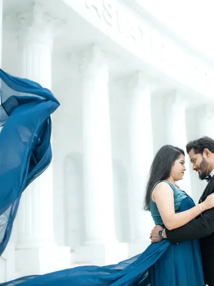 A dramatic, cinematic shot featuring a flowing blue gown against the grand, all-white architecture of a studio set. This style is perfect for couples who dream of a filmi, larger-than-life pre-wedding album.