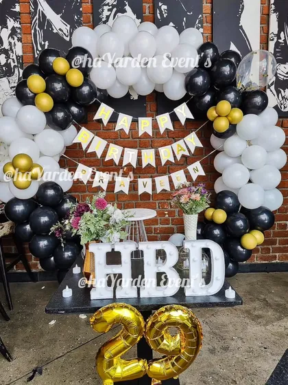 A fun and festive 29th birthday party. The black, white, and gold balloon arch is paired with large 'HBD' marquee letters and foil '29' balloons.