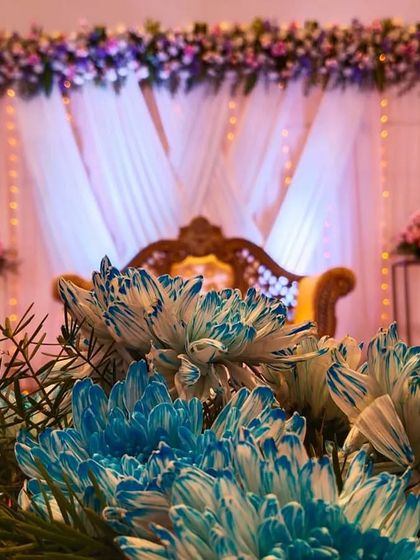 A close-up artistic shot of the reception decor, focusing on a bouquet of blue and white dyed chrysanthemums in the foreground with the beautifully lit stage blurred in the background.