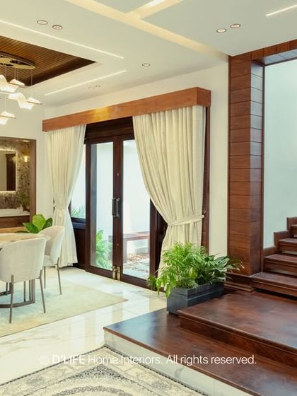 An elegant dining room in a contemporary Munnar home, featuring a stunning modern chandelier over a light-toned table, with a view into the sleek black kitchen.