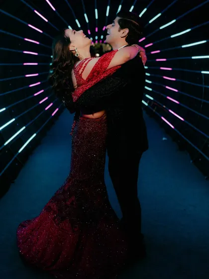 A glamorous engagement photo of a couple sharing a joyful moment. Her sparkling red gown and his classic black suit create a dramatic and celebratory feel, enhanced by the tunnel of neon lights.