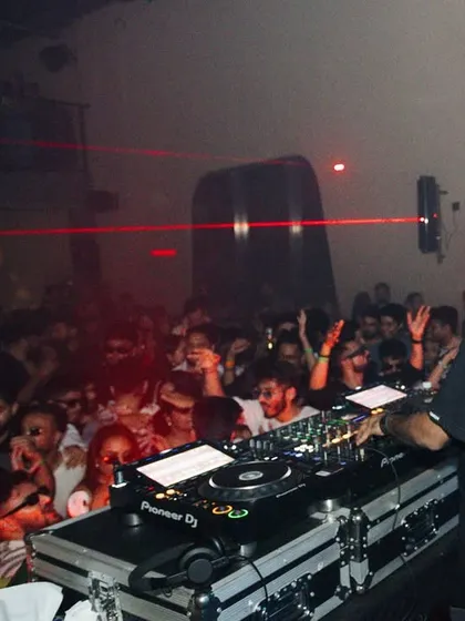 A view from behind the decks, showing the packed crowd and laser beams cutting through the darkness.