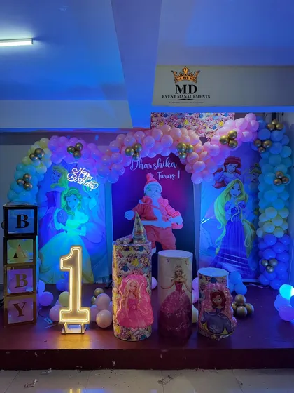 A grand Disney Princess theme for a first birthday party. The stage is decorated with character backdrops of Cinderella and Rapunzel, a balloon arch, 'BABY' blocks, and a light-up number '1'.