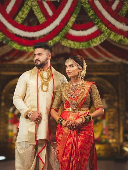 The couple standing together, framed by the rich textures of the temple mandap and the vibrant red and white floral decorations hanging from the ceiling.