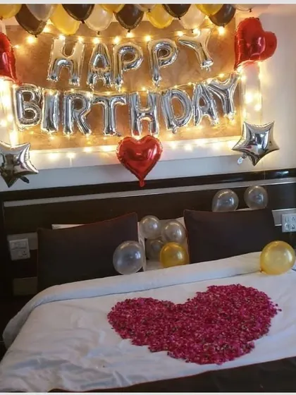 A romantic birthday surprise with a heart made of rose petals on the bed. The wall is decorated with 'Happy Birthday' foil letters and star-shaped balloons.