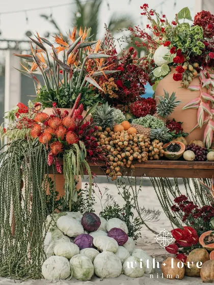 An abundant display of tropical fruits and flowers, including coconuts, cabbage, and pineapples, creating a rich and textured installation on the beach.