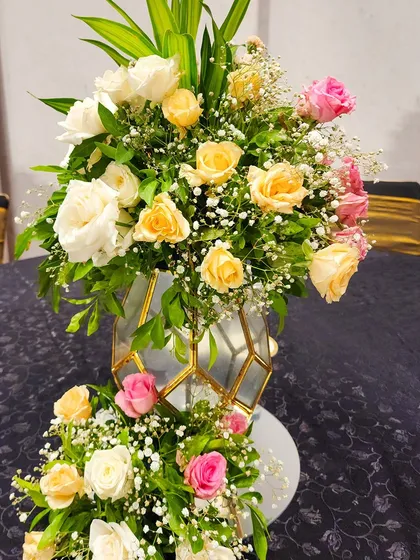 A beautiful floral arrangement in a geometric gold and glass vase. The bouquet features a soft palette of yellow, white, and pink roses, perfect for an elegant reception or gala dinner.