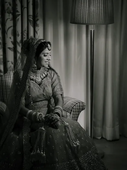 A timeless black and white bridal portrait. The dramatic lighting from the side creates a classic, elegant mood, highlighting the bride's profile and the texture of her lehenga.