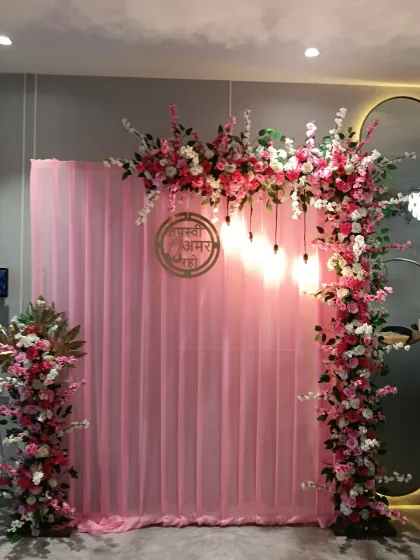 An asymmetrical floral arch in pink and white against a sheer pink curtain. This decoration for a Tapasvi event includes a custom golden sign and hanging lights.
