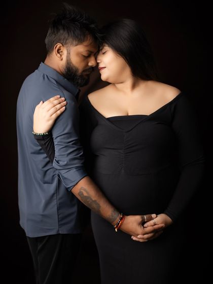 An intimate close-up of a couple in a dark studio setting. Dressed in dark, coordinating outfits, they share a tender forehead-to-forehead moment, their hands joined over the baby bump.