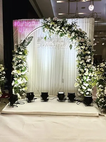 A beautiful photo backdrop for an engagement, featuring a circular arch of white flowers and a custom neon sign with the couple's hashtag, creating a perfect spot for memories.