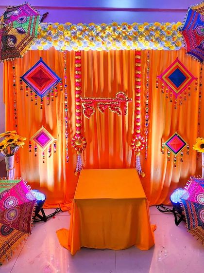 A traditional and budget-friendly Haldi setup. This design uses orange drapes, colorful Rajasthani umbrellas, and hanging elements to create a festive and bright atmosphere.