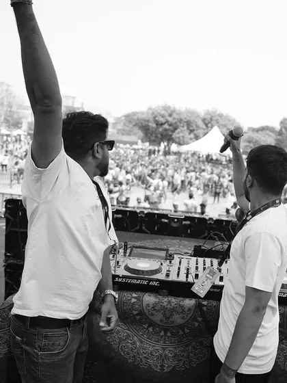 A black and white shot from the Holi festival stage, raising a hand to the massive crowd. The energy exchange is powerful.