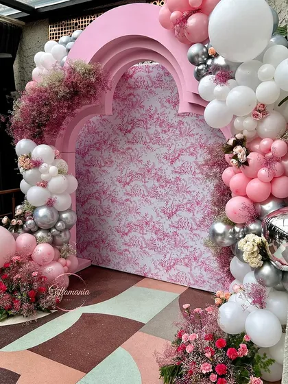 This Dior-inspired 40th birthday party backdrop features a custom-printed panel in a signature pattern, framed by an elegant pink arch. The look is completed with a cascading garland of pink, white, and silver balloons mixed with fresh floral accents.