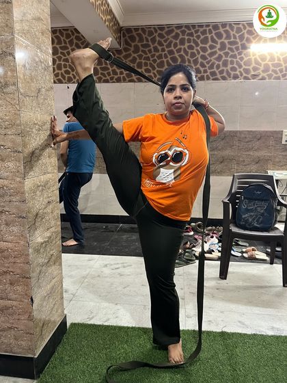 This student is practicing Trivikram Asana, or Standing Splits, using a belt and the wall for support. This method helps to safely work on extreme flexibility while maintaining balance.