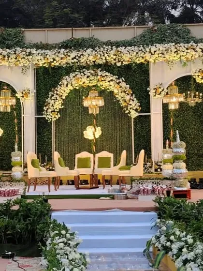 I designed this elegant outdoor reception stage with a backdrop of lush greenery and white floral arches. The setup includes beautiful chandeliers and comfortable seating, creating a sophisticated and romantic ambiance for the evening.