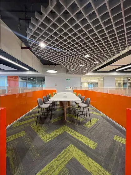 A long collaborative table set against a vibrant orange partition wall, designed for quick team huddles. The grid ceiling and patterned carpet define this zone within the larger workspace, demonstrating our use of design to guide function.