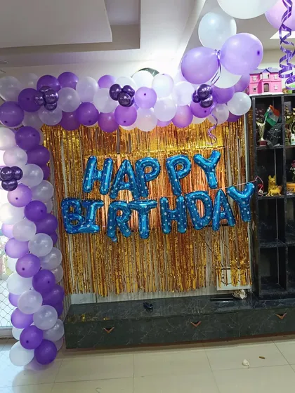 A corner balloon arch in purple and white against a gold tinsel curtain, with blue 'Happy Birthday' foil letters.