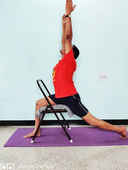 Veerabhadrasana 1 (Warrior 1) with the front thigh supported by a chair. This modification helps in squaring the hips and extending the spine upwards without straining the lower back.