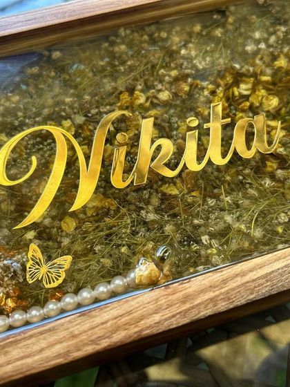 A close-up of a wooden frame nameplate made with preserved varmala flowers. The gold lettering and butterfly charm add a beautiful finishing touch.