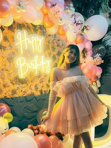 A guest posing at a birthday party with a beautiful setup featuring a "Happy Birthday" neon sign and a large, illuminated letter "D". The pink and white balloon decor adds a soft, celebratory feel.