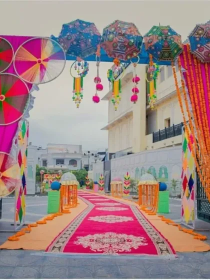 A grand and colorful entrance for a Rajasthani-themed Mehendi event. The arch is decorated with pinwheels and umbrellas, leading to a red carpet walkway.