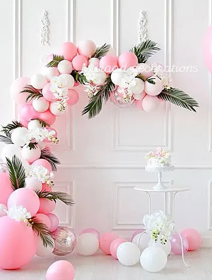 An elegant pink and white balloon arch decorated with faux flowers and palm leaves. This setup against a clean white wall is perfect for a sophisticated garden party or bridal shower.