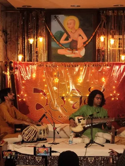 The stage is set under the watchful gaze of Saraswati, the goddess of music. The atmosphere is one of reverence and artistry.