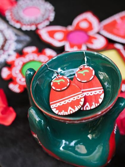 A close-up of vibrant, hand-painted wooden earrings, displayed in a quirky hand-shaped ceramic bowl.