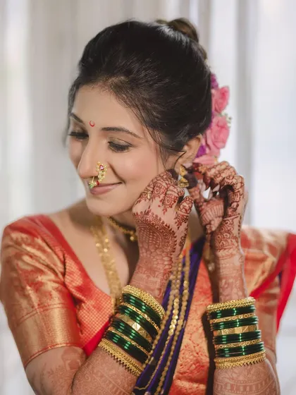 A close-up of the bride Aditi as she puts on her earrings. This shot highlights the detailed henna and the beautiful green bangles, essential parts of the Marathi bridal attire.