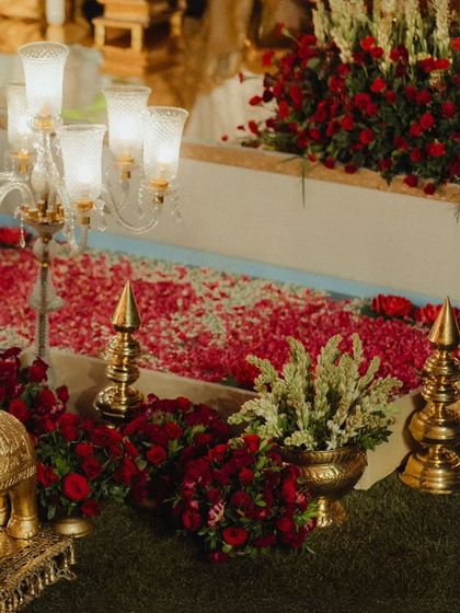 A close-up of the floral and brass decor lining the aisle. The path is covered in a carpet of red and pink rose petals, flanked by candelabras and traditional brass finials, creating a truly royal passage.