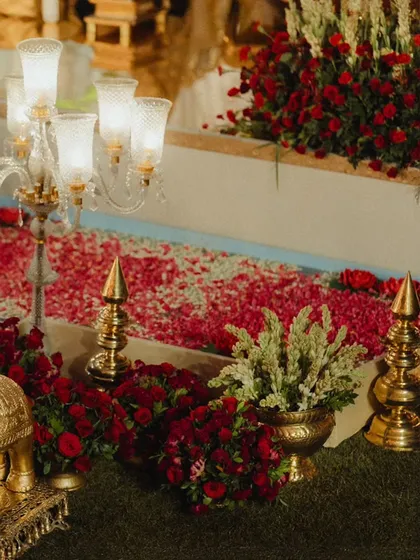 A close-up of the floral and brass decor lining the aisle. The path is covered in a carpet of red and pink rose petals, flanked by candelabras and traditional brass finials, creating a truly royal passage.