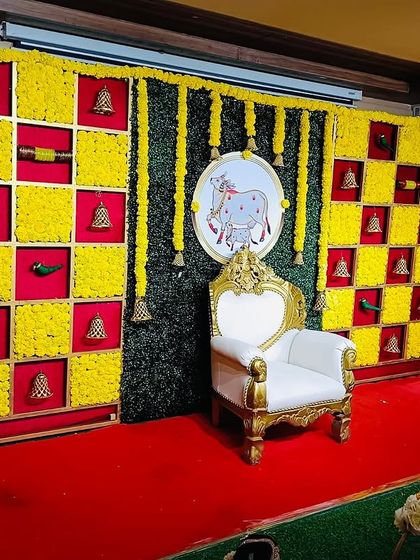 A classic Seemantham backdrop with a checkerboard of yellow marigolds and red fabric. The design is accented with small bells and parrot motifs, with a central elephant painting for good luck.