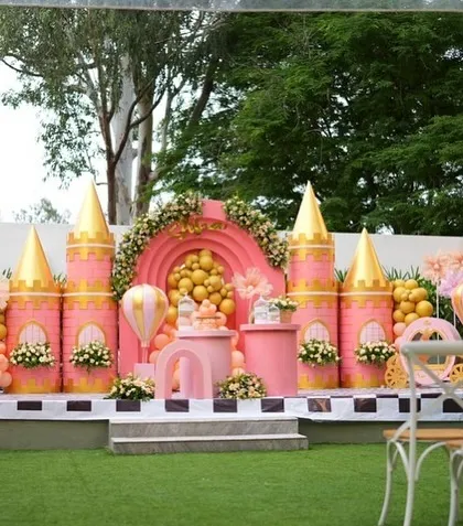 A wider view of Soha's pink and gold Disney-style castle, set up outdoors on a green lawn. The setup includes seating for guests, making it a perfect stage for a birthday ceremony.