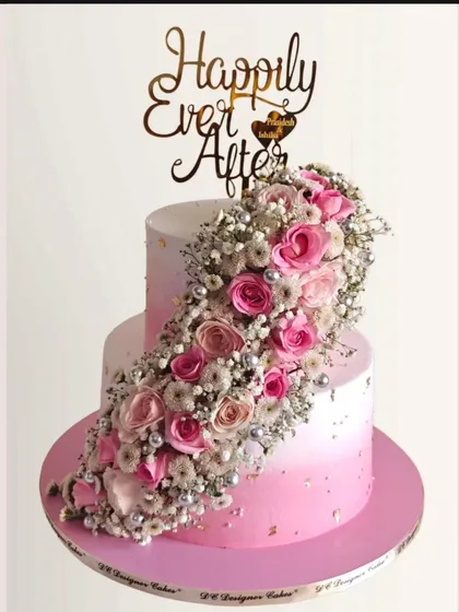 A romantic two-tier wedding cake with a cascade of fresh pink roses and baby's breath. The pink ombre effect on the whipped cream adds a soft, modern touch.