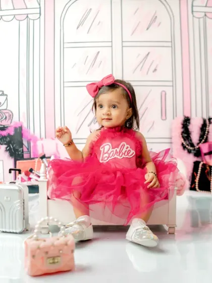 Welcome to Barbie's World! This toddler is perfectly styled for her first birthday shoot with a vibrant pink dress and bow, sitting on a miniature bed in a chic, illustrated boutique setting.
