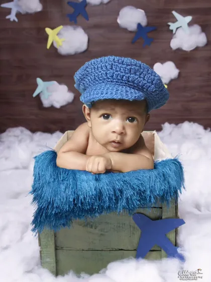 A baby in a blue cap peeking out of a crate, with a background of clouds and airplanes.