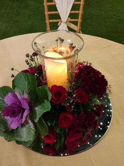 This centerpiece combines deep red roses and carnations with a unique ornamental cabbage. The arrangement sits on a mirrored base, surrounding a candle in a glass hurricane lamp for a touch of warmth and color.