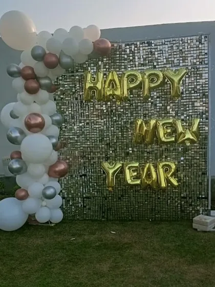 A festive 'Happy New Year' photo backdrop for a corporate party or event. A silver sequin wall is paired with a simple balloon garland and gold foil letters.