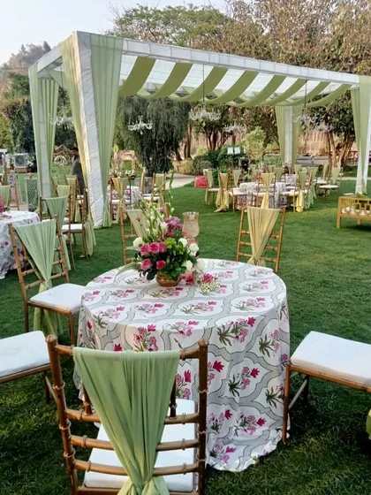 An elegant outdoor seating arrangement for a day wedding. The tables are covered with floral-printed cloths, and the chairs are decorated with sage green drapes.