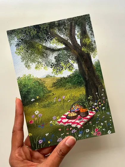 This gouache painting captures the perfect cottagecore aesthetic with a picnic scene under a large tree in a field of wildflowers. It’s a piece of art that feels like a warm, happy memory.
