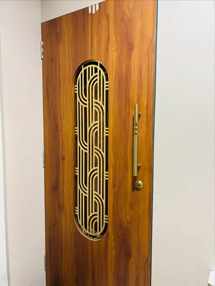 A close-up of a wooden safety door with an elegant, interwoven brass grill design. The long brass handle complements the grill and adds a touch of luxury.