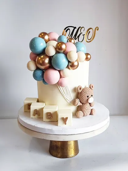 A classic and beautiful baby shower cake for a lovely couple. This design features a teddy bear holding a cascade of pink, blue, and gold chocolate balloons, with edible 'BABY' blocks.