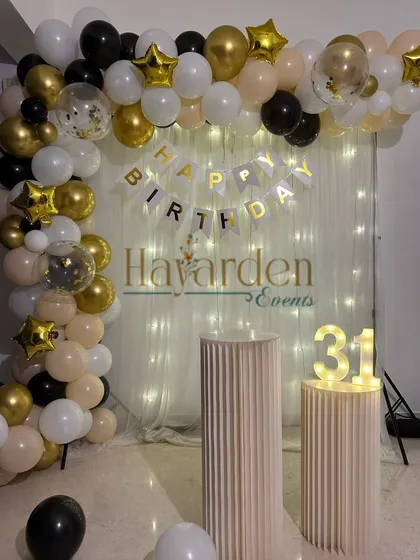 A stylish 31st birthday setup. The decor features a balloon arch in neutral tones with black and gold accents, a fairy light curtain, and a small light-up '31'.