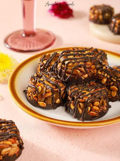 A close-up of the delicious nut clusters, showing the glossy chocolate and crunchy texture. The soft pink background gives it a sweet and celebratory feel.